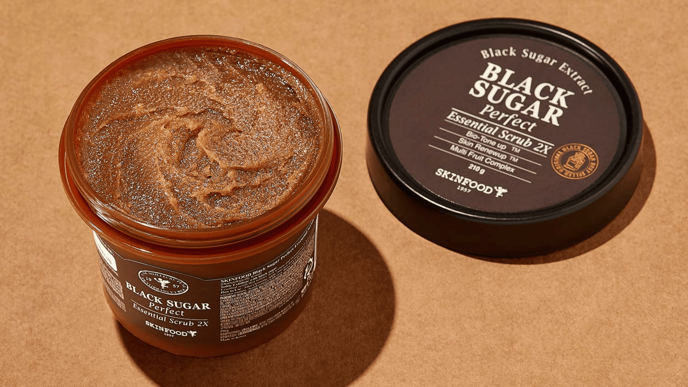 En öppen burk med Skinfood Black Sugar Perfect Essential Scrub 2X, bredvid dess svarta lock, på en brun bakgrund.