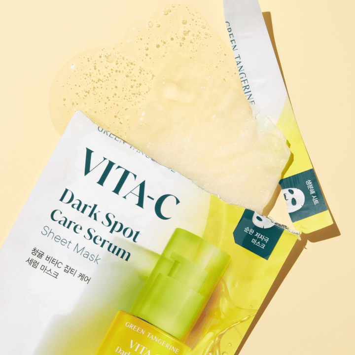 Öppnad förpackning av VITA-C Dark Spot Care Serum Sheet Mask med en ansiktsmask ovanpå och en grön serumflaska, alla mot en gul bakgrund.